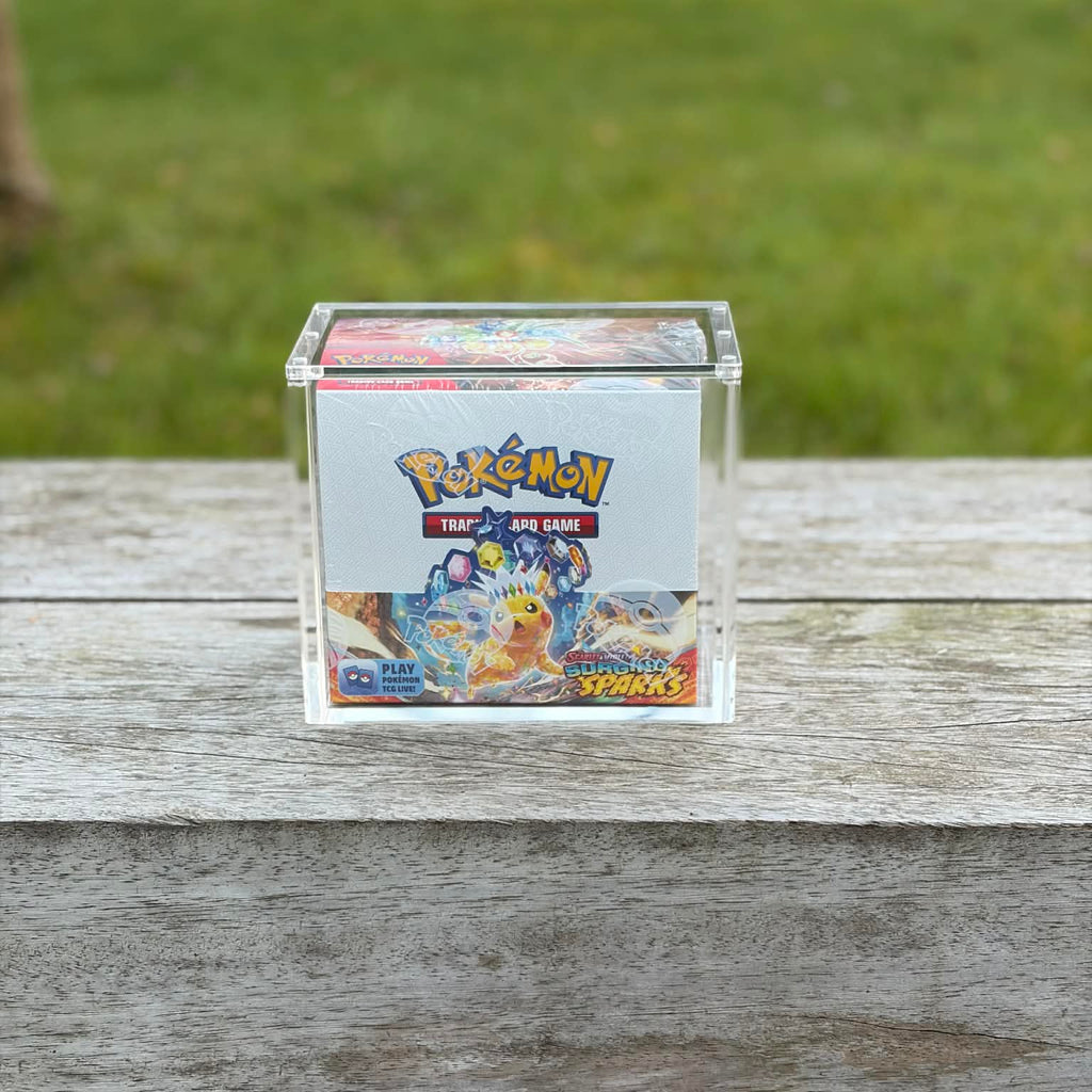 Pokémon trading card game box in a clear acrylic display case on a wooden surface with grass in the background.