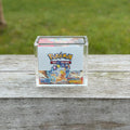 Pokémon trading card game box in a clear acrylic display case on a wooden surface with grass in the background.